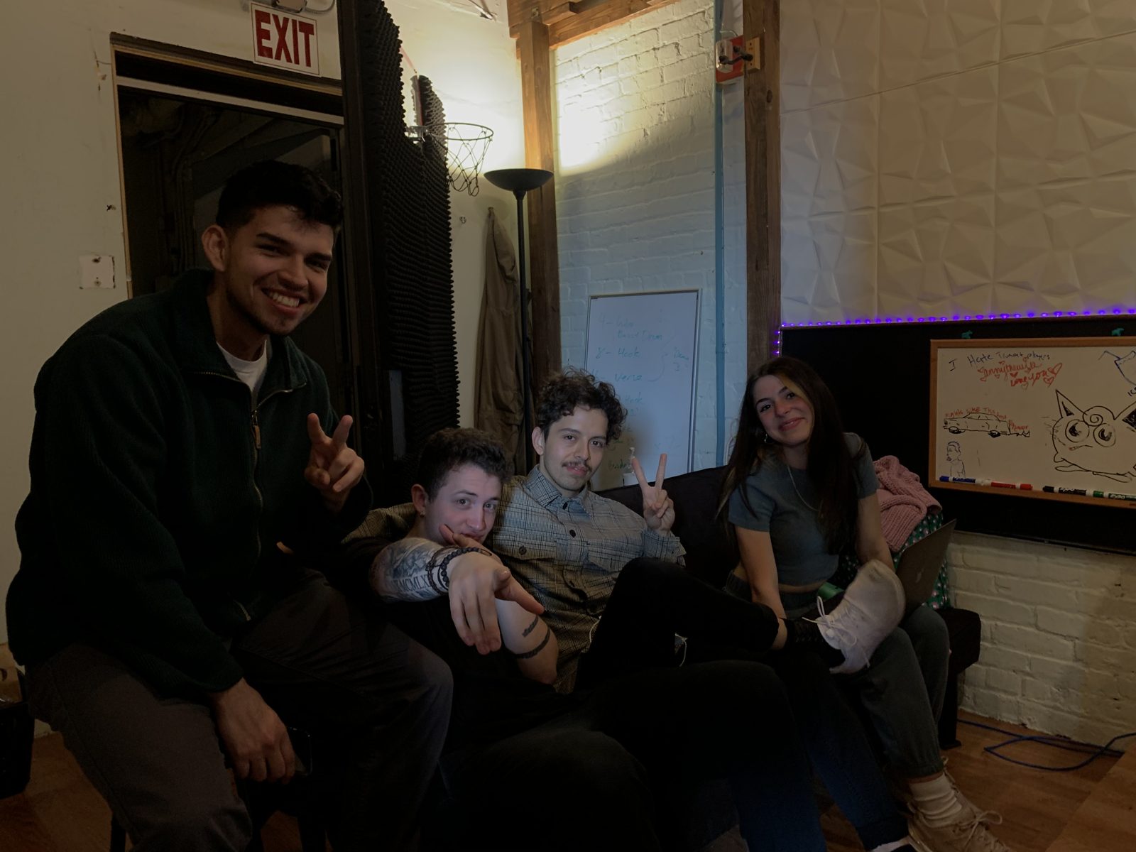 Four people on a couch in a studio lounge with a brick wall and whiteboards, two throwing peace signs.