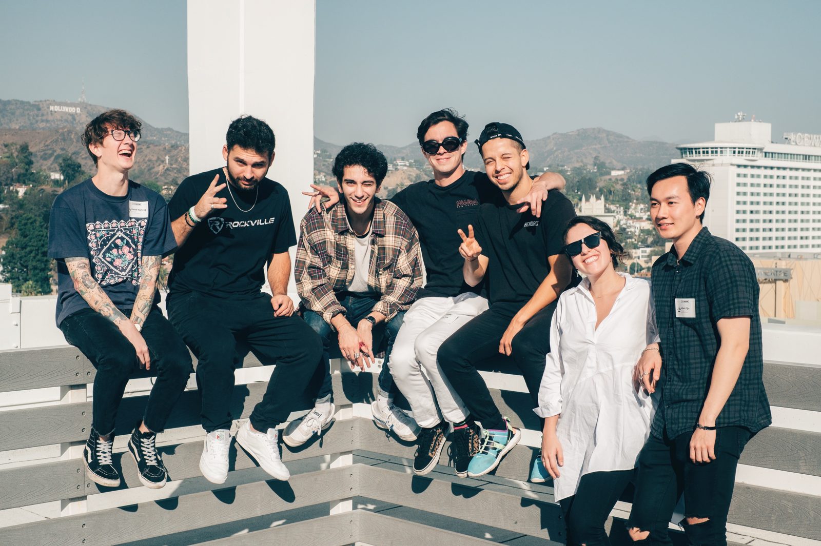 Seven people seated on a rooftop railing in Los Angeles, the Hollywood sign on the hills behind them.